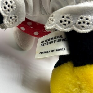 May include: A plush toy featuring a white dress with eyelet lace trim, a red polka dot bow, and a yellow foot. The tag reads "ALL NEW MATERIAL POLYESTER STUFFING PRODUCT OF KOREA."