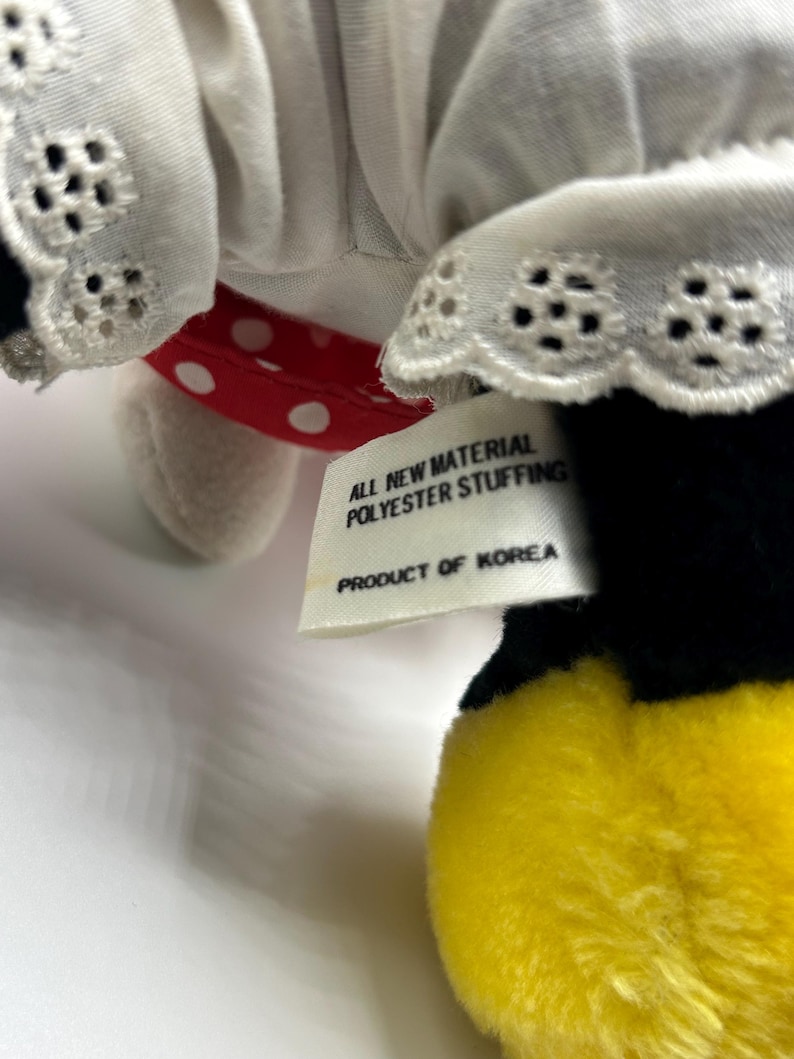 May include: A plush toy featuring a white dress with eyelet lace trim, a red polka dot bow, and a yellow foot. The tag reads "ALL NEW MATERIAL POLYESTER STUFFING PRODUCT OF KOREA."
