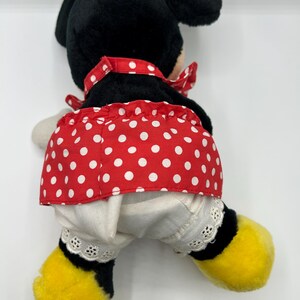 May include: A plush Minnie Mouse doll, viewed from the back, features a black head and ears, a red polka-dot skirt, and white bloomers with lace trim. The doll has yellow feet and a red bow.