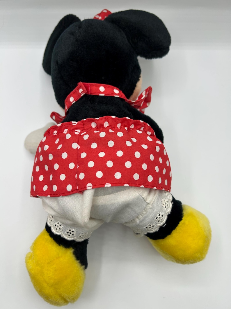 May include: A plush Minnie Mouse doll, viewed from the back, features a black head and ears, a red polka-dot skirt, and white bloomers with lace trim. The doll has yellow feet and a red bow.