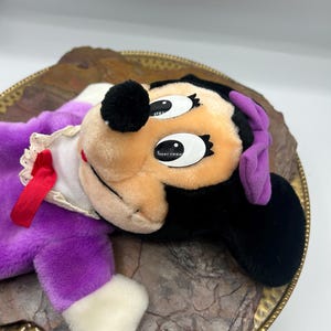 May include: A vintage Minnie Mouse plush doll, featuring a black head with large eyes, a tan face, and a red bow. The doll is dressed in a purple dress with white trim and a red ribbon. The plush is resting on a decorative gold-rimmed plate.