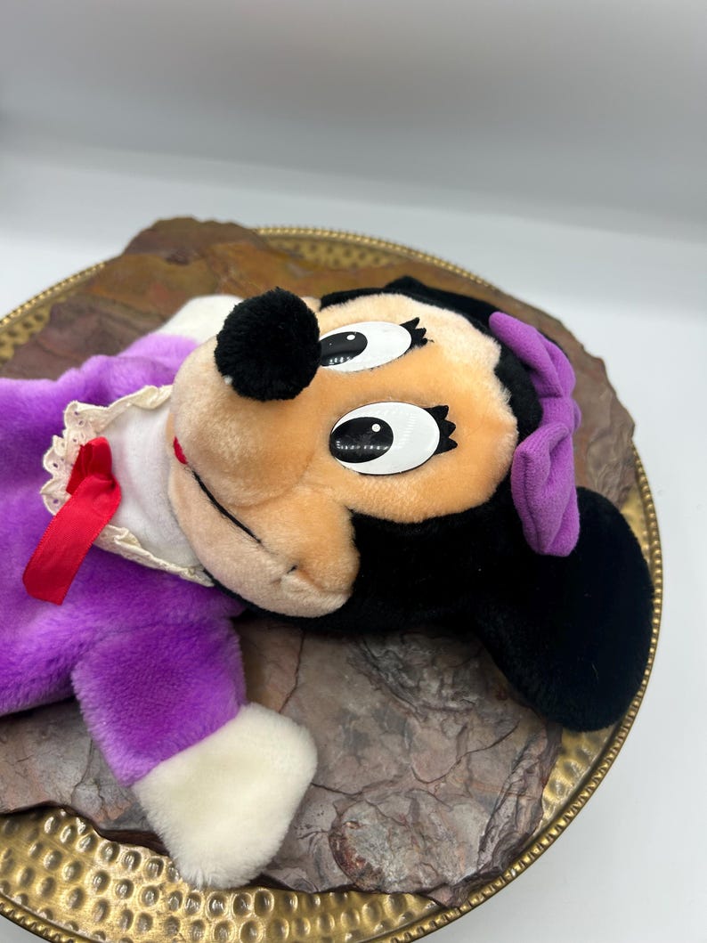 May include: A vintage Minnie Mouse plush doll, featuring a black head with large eyes, a tan face, and a red bow. The doll is dressed in a purple dress with white trim and a red ribbon. The plush is resting on a decorative gold-rimmed plate.