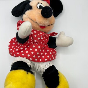 May include: A plush Minnie Mouse doll featuring a red and white polka dot dress and bow. The doll has black ears, a black nose, and yellow feet. The doll's arms and hands are white.