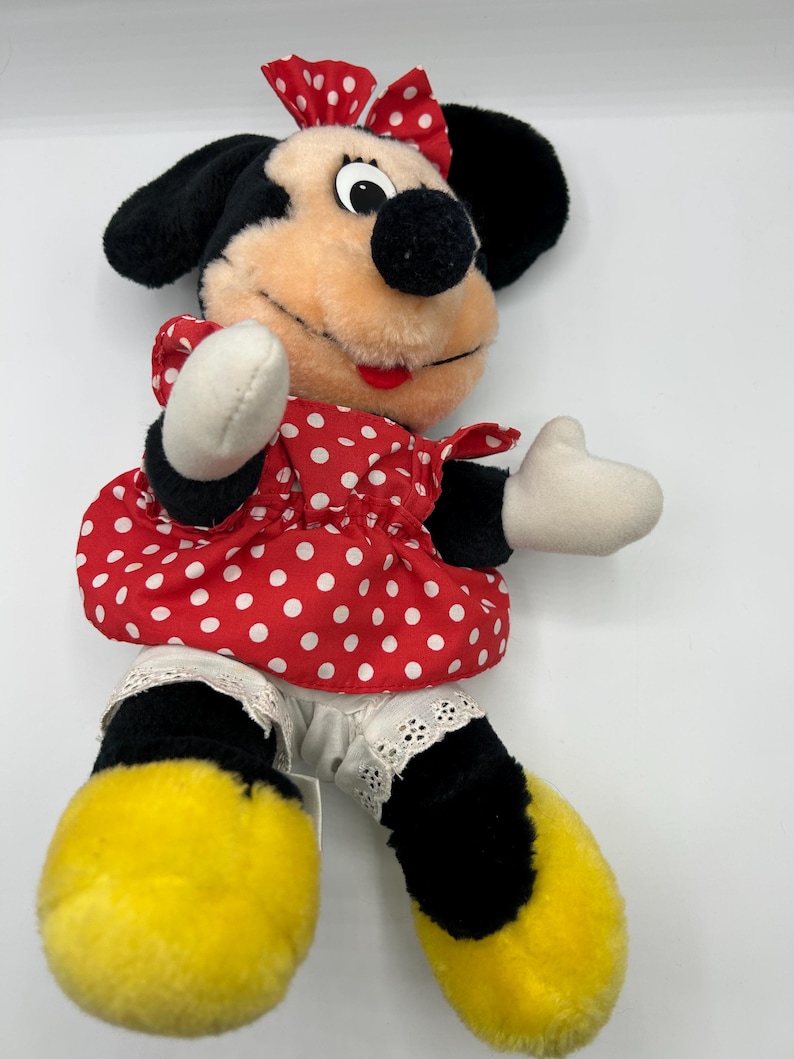 May include: A plush Minnie Mouse doll featuring a red and white polka dot dress and bow. The doll has black ears, a black nose, and yellow feet. The doll's arms and hands are white.