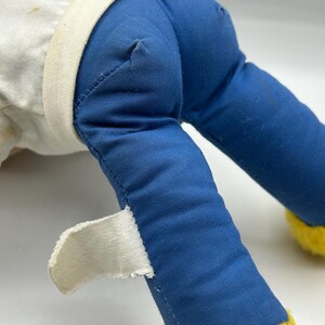 Peut inclure: Gros plan sur les jambes et le torse d'une poup&eacute;e en peluche vintage. La poup&eacute;e porte un pantalon en tissu bleu, une chemise blanche avec un col et des pieds jaunes et flous. Une &eacute;tiquette blanche est attach&eacute;e &agrave; la jambe de la poup&eacute;e.