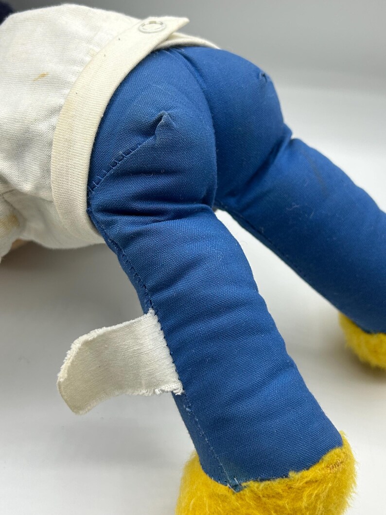 Peut inclure: Gros plan sur les jambes et le torse d'une poup&eacute;e en peluche vintage. La poup&eacute;e porte un pantalon en tissu bleu, une chemise blanche avec un col et des pieds jaunes et flous. Une &eacute;tiquette blanche est attach&eacute;e &agrave; la jambe de la poup&eacute;e.