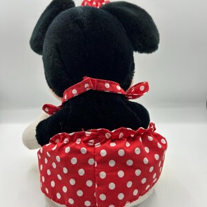 May include: A plush toy of a cartoon character, viewed from the back. The toy has black fur, large black ears, and a red bow with white polka dots. It wears a red skirt with white polka dots and a red collar.