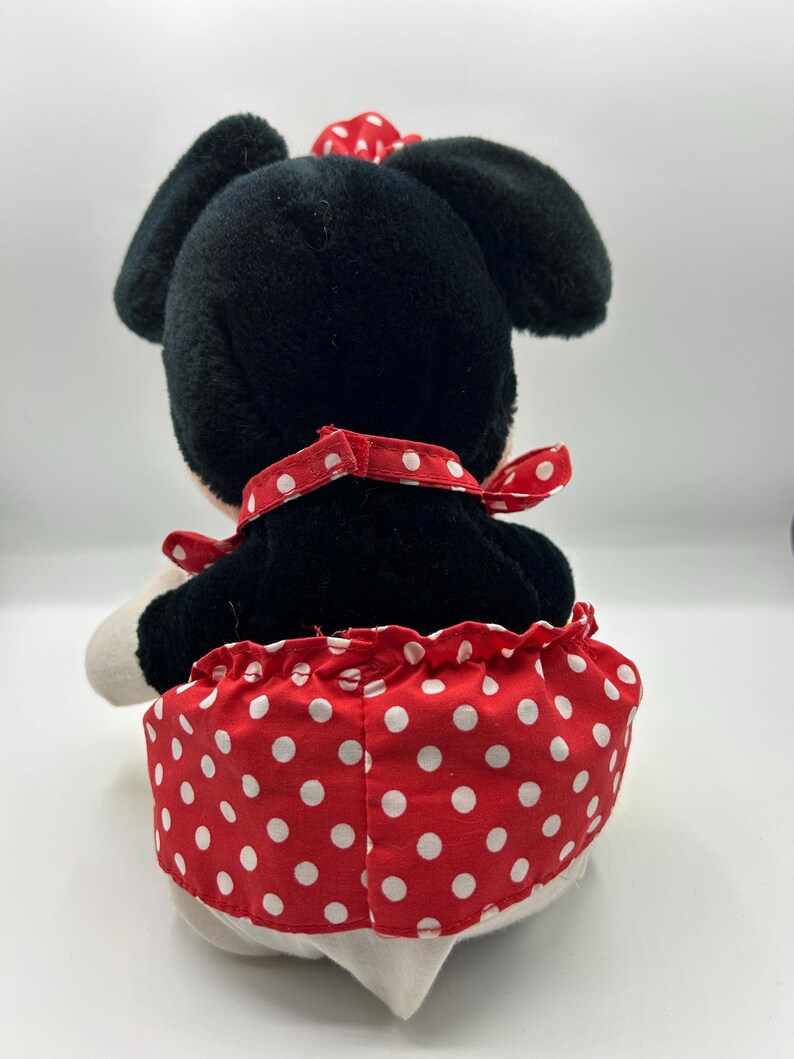 May include: A plush toy of a cartoon character, viewed from the back. The toy has black fur, large black ears, and a red bow with white polka dots. It wears a red skirt with white polka dots and a red collar.