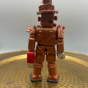 K&ouml;nnte beinhalten: Ein bronzefarbener Spielzeugroboter mit einer hutartigen Struktur auf dem Kopf. Der Roboter hat eine rote Hand und eine silberne Hand. Der Roboter steht auf einem goldfarbenen Tablett.