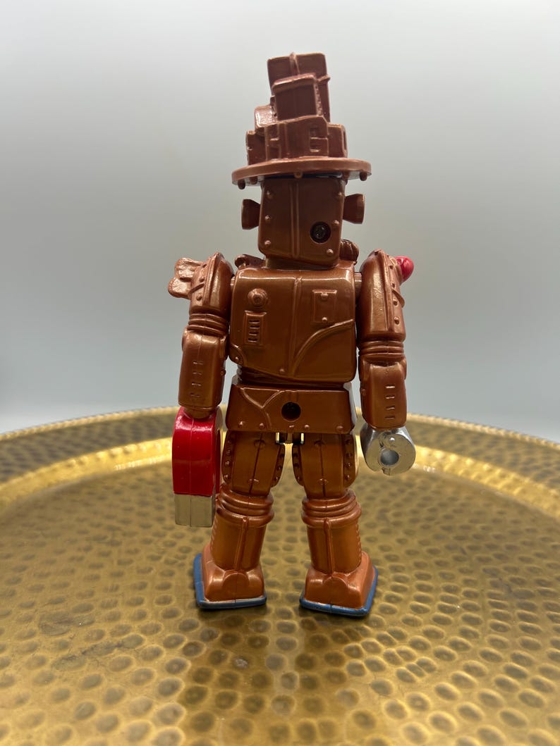 K&ouml;nnte beinhalten: Ein bronzefarbener Spielzeugroboter mit einer hutartigen Struktur auf dem Kopf. Der Roboter hat eine rote Hand und eine silberne Hand. Der Roboter steht auf einem goldfarbenen Tablett.