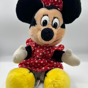 May include: A plush Minnie Mouse doll. The doll has black ears, a red and white polka dot bow, and a matching dress. Her large yellow feet and white gloves are visible. The doll is sitting and has a smiling expression.