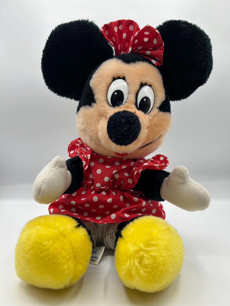 May include: A plush Minnie Mouse doll. The doll has black ears, a red and white polka dot bow, and a matching dress. Her large yellow feet and white gloves are visible. The doll is sitting and has a smiling expression.