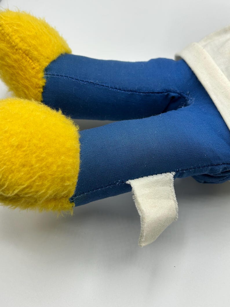 Peut inclure: Gros plan sur les jambes et une partie du torse d'une peluche. Les jambes sont bleues avec des pieds jaunes et duveteux. Une &eacute;tiquette blanche est attach&eacute;e au tissu bleu. Le torse est blanc.
