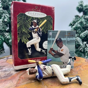 以下が含まれることがあります： 野球のユニフォームを着たハンク・アーロンが描かれた赤いHallmark Keepsake Ornamentの箱。野球のユニフォームを着たハンク・アーロンのフィギュアが横たわっています。ハンク・アーロンの野球カードも存在します。