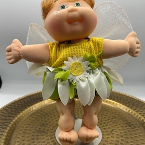 Puede incluir: Una muñeca con cabello castaño rojizo y ojos verdes, que viste un top amarillo texturizado y una falda hecha de pétalos de margarita blancos con hojas verdes. La muñeca tiene alas blancas translúcidas y una pequeña margarita en la mejilla.