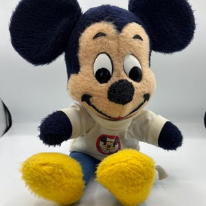 Peut inclure: Peluche vintage de Mickey Mouse. La peluche pr&eacute;sente un visage beige, un nez noir et de grandes oreilles noires. Mickey porte une chemise blanche avec un graphisme circulaire et un pantalon bleu. Ses grands pieds sont jaunes.