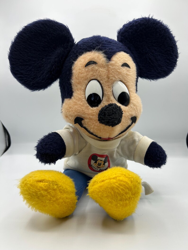 Peut inclure: Peluche vintage de Mickey Mouse. La peluche pr&eacute;sente un visage beige, un nez noir et de grandes oreilles noires. Mickey porte une chemise blanche avec un graphisme circulaire et un pantalon bleu. Ses grands pieds sont jaunes.