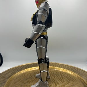 K&ouml;nnte beinhalten: Seitenansicht einer detaillierten Actionfigur. Die Figur ist haupts&auml;chlich grau, schwarz und goldfarben, mit roten Akzenten auf dem Helm. Die Figur hat ein schlankes, gepanzertes Design mit beweglichen Gelenken und einer dynamischen Pose. Die Figur steht auf einer goldfarbenen, strukturierten Oberfl&auml;che.