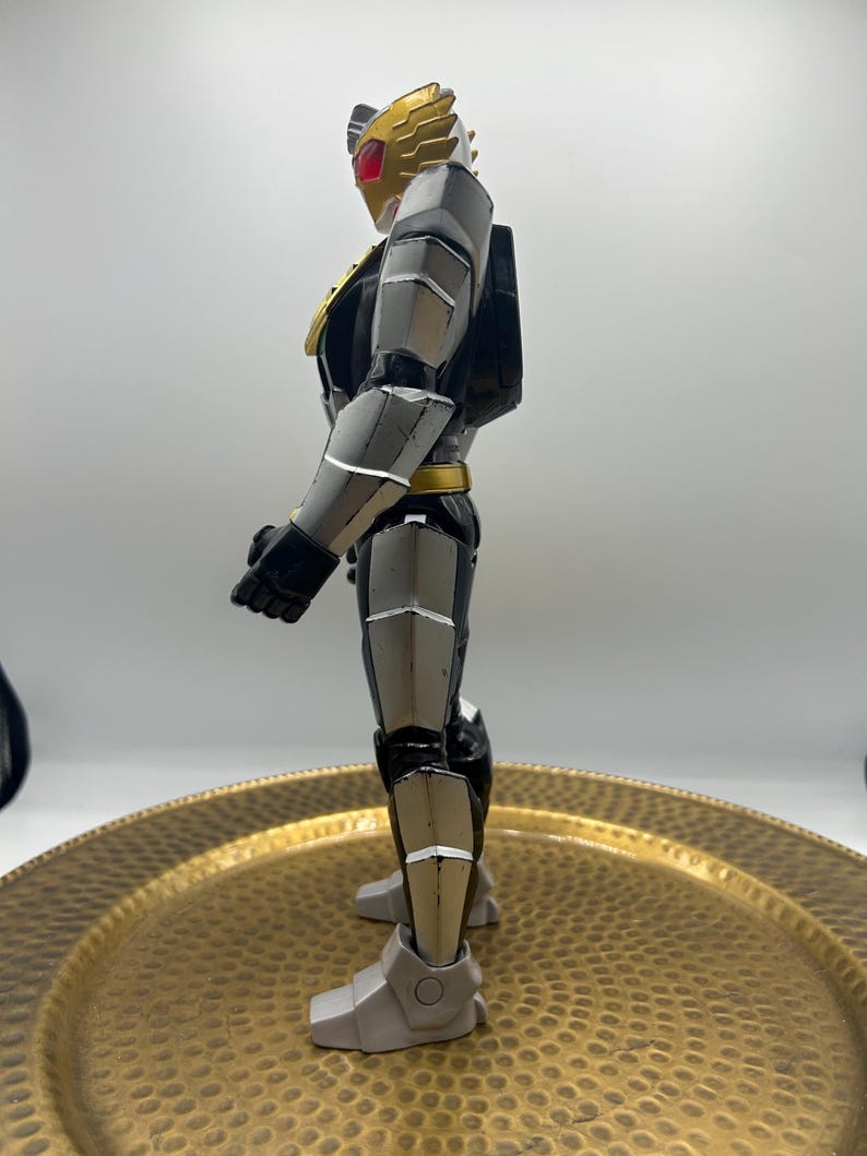 K&ouml;nnte beinhalten: Seitenansicht einer detaillierten Actionfigur. Die Figur ist haupts&auml;chlich grau, schwarz und goldfarben, mit roten Akzenten auf dem Helm. Die Figur hat ein schlankes, gepanzertes Design mit beweglichen Gelenken und einer dynamischen Pose. Die Figur steht auf einer goldfarbenen, strukturierten Oberfl&auml;che.