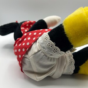 May include: A plush toy of a cartoon character, featuring black ears, a red dress with white polka dots, and white bloomers with lace trim. The character's yellow feet are visible, and the toy is lying on its side.
