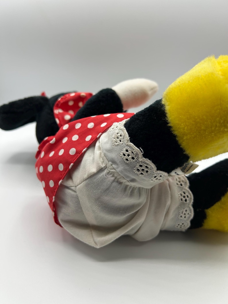 May include: A plush toy of a cartoon character, featuring black ears, a red dress with white polka dots, and white bloomers with lace trim. The character's yellow feet are visible, and the toy is lying on its side.