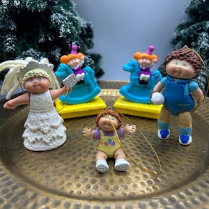 Può includere: Una collezione di cinque ornamenti Cabbage Patch Kids che include una bambola con un vestito bianco, due bambole su cavalli a dondolo blu con basi gialle, una bambola con salopette blu che tiene una palla e una piccola bambola con salopette gialla.
