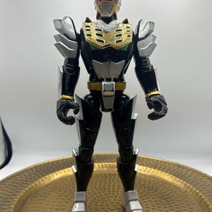 K&ouml;nnte beinhalten: Eine schwarz-silberne Actionfigur mit goldenen und roten Akzenten. Die Figur, mit detailliertem Helm und R&uuml;stung, steht auf einem goldfarbenen Zierteller. Das Design der Figur erinnert an einen Superhelden oder Roboter.