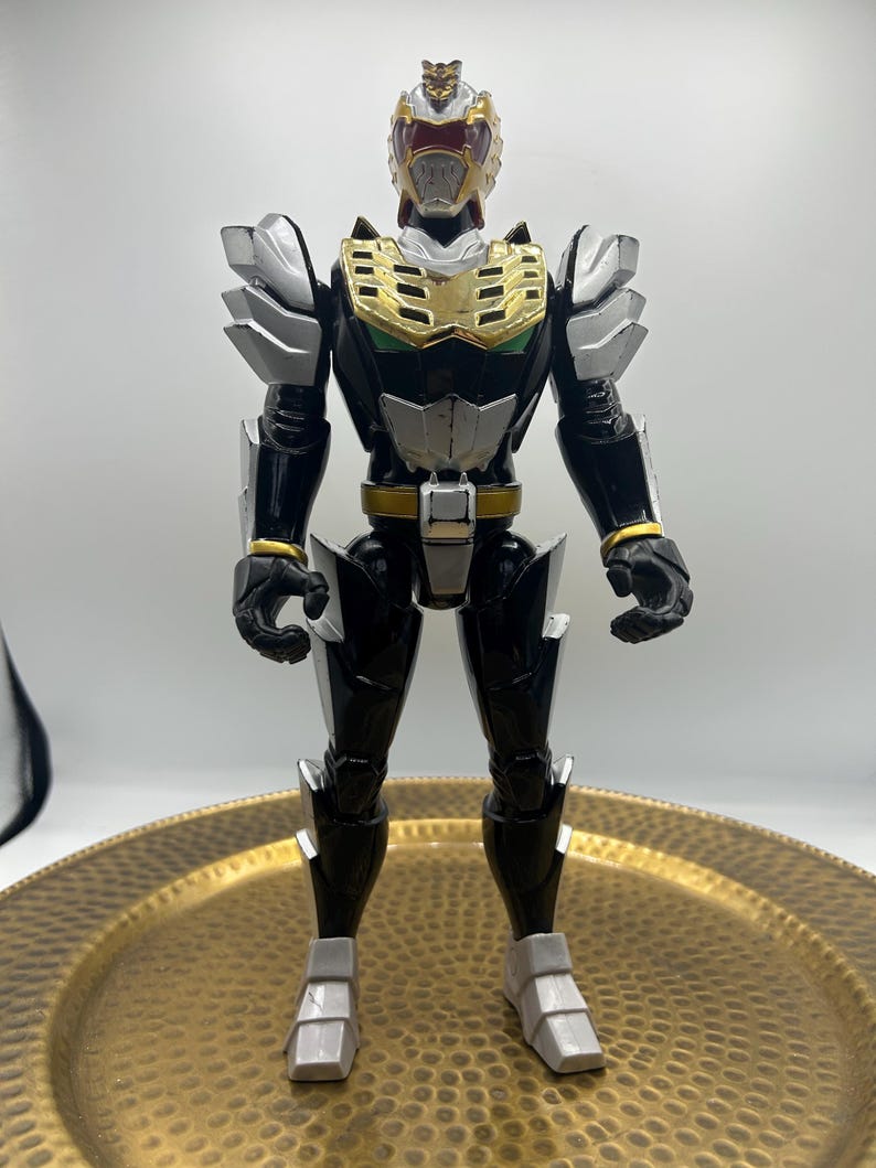 K&ouml;nnte beinhalten: Eine schwarz-silberne Actionfigur mit goldenen und roten Akzenten. Die Figur, mit detailliertem Helm und R&uuml;stung, steht auf einem goldfarbenen Zierteller. Das Design der Figur erinnert an einen Superhelden oder Roboter.