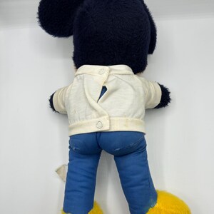 Peut inclure: Peluche vintage de Mickey Mouse, vue de dos. La peluche pr&eacute;sente une t&ecirc;te bleu fonc&eacute; avec de grandes oreilles, une veste blanche, un pantalon bleu et des chaussures jaunes. La veste a deux boutons. Le jouet est sur fond blanc.