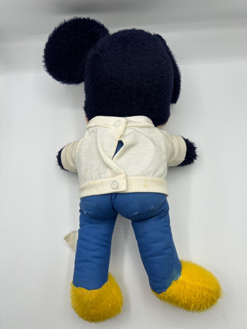 Peut inclure: Peluche vintage de Mickey Mouse, vue de dos. La peluche pr&eacute;sente une t&ecirc;te bleu fonc&eacute; avec de grandes oreilles, une veste blanche, un pantalon bleu et des chaussures jaunes. La veste a deux boutons. Le jouet est sur fond blanc.
