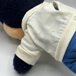 Peut inclure: Une peluche avec une t&ecirc;te bleu fonc&eacute; et un visage beige. Le jouet porte une chemise blanche et un pantalon bleu. La chemise a un col boutonn&eacute; et des manches longues. Les pieds du jouet sont bleu fonc&eacute;.