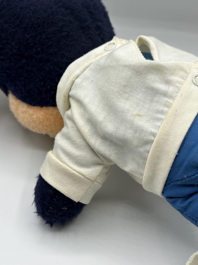 Peut inclure: Une peluche avec une t&ecirc;te bleu fonc&eacute; et un visage beige. Le jouet porte une chemise blanche et un pantalon bleu. La chemise a un col boutonn&eacute; et des manches longues. Les pieds du jouet sont bleu fonc&eacute;.