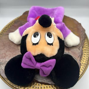 May include: A plush toy featuring a cartoon character with a black head, ears, and body, a peach-colored face, and large, expressive eyes. It has a purple bow tie and a purple outfit. The toy is lying on a decorative plate.