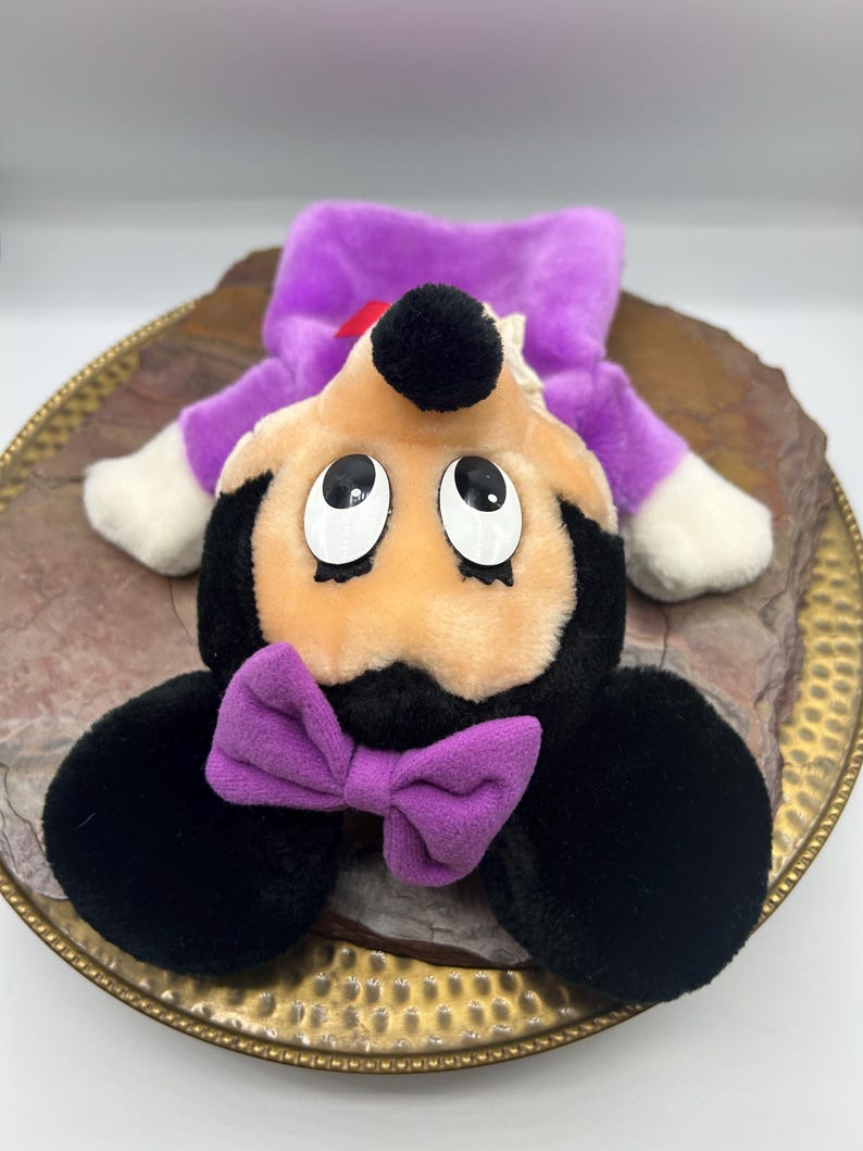 May include: A plush toy featuring a cartoon character with a black head, ears, and body, a peach-colored face, and large, expressive eyes. It has a purple bow tie and a purple outfit. The toy is lying on a decorative plate.