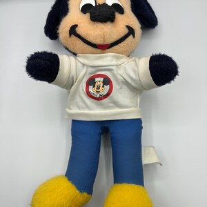 Peut inclure: Une poup&eacute;e en peluche vintage de Mickey Mouse. La poup&eacute;e a un visage bronz&eacute;, des oreilles et des bras noirs, et un pantalon bleu. Elle porte un t-shirt blanc avec un logo Mickey Mouse Club et des pieds jaunes. Un objet de collection Disney classique.