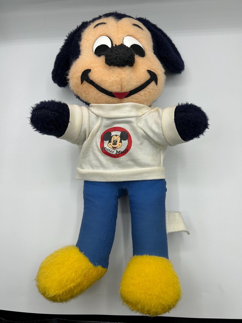 Peut inclure: Une poup&eacute;e en peluche vintage de Mickey Mouse. La poup&eacute;e a un visage bronz&eacute;, des oreilles et des bras noirs, et un pantalon bleu. Elle porte un t-shirt blanc avec un logo Mickey Mouse Club et des pieds jaunes. Un objet de collection Disney classique.