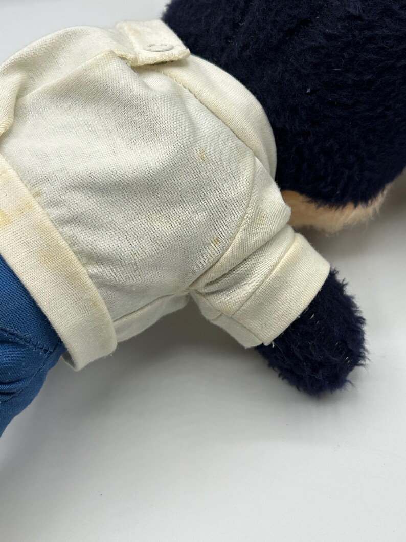 Peut inclure: Un ours en peluche vintage avec une chemise blanche et un pantalon bleu. L'ours a une fourrure bleu fonc&eacute; et un museau beige clair. La chemise a une fermeture &agrave; bouton et les bras et les jambes de l'ours sont visibles.