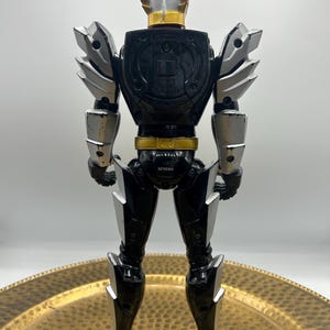 K&ouml;nnte beinhalten: Eine schwarz-silberne Actionfigur mit goldenen Akzenten. Die Figur hat einen Helm mit silberner Oberseite und goldenem Rand. Schultern und Arme haben silberne R&uuml;stungen. Die Figur steht auf einer goldfarbenen Oberfl&auml;che.