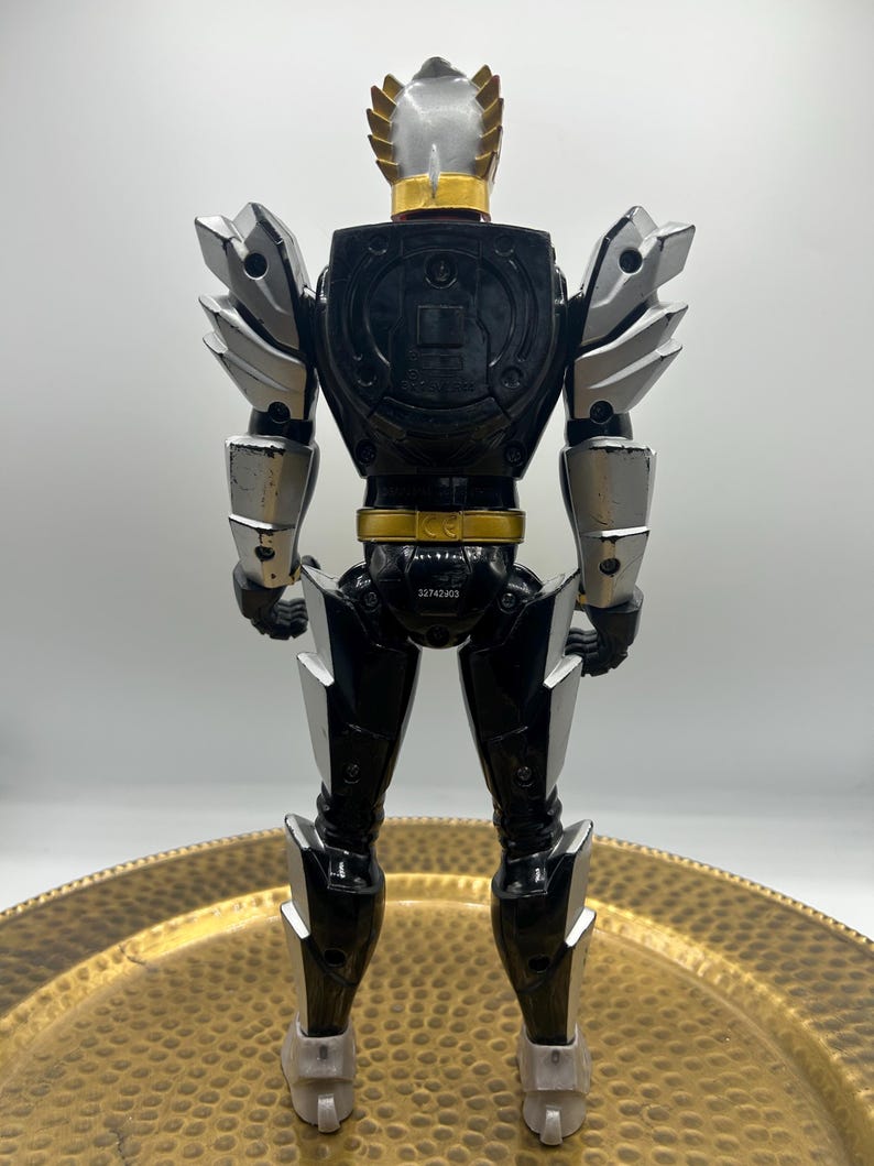 K&ouml;nnte beinhalten: Eine schwarz-silberne Actionfigur mit goldenen Akzenten. Die Figur hat einen Helm mit silberner Oberseite und goldenem Rand. Schultern und Arme haben silberne R&uuml;stungen. Die Figur steht auf einer goldfarbenen Oberfl&auml;che.