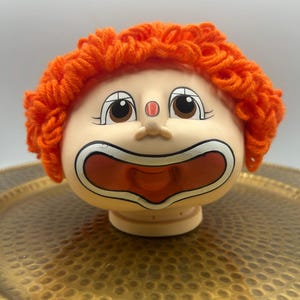 May include: A plastic doll head with bright orange yarn hair, featuring a painted face with large brown eyes, a small red nose, and a wide, open mouth with white and black outlines. The doll head has a smooth, light-toned complexion.