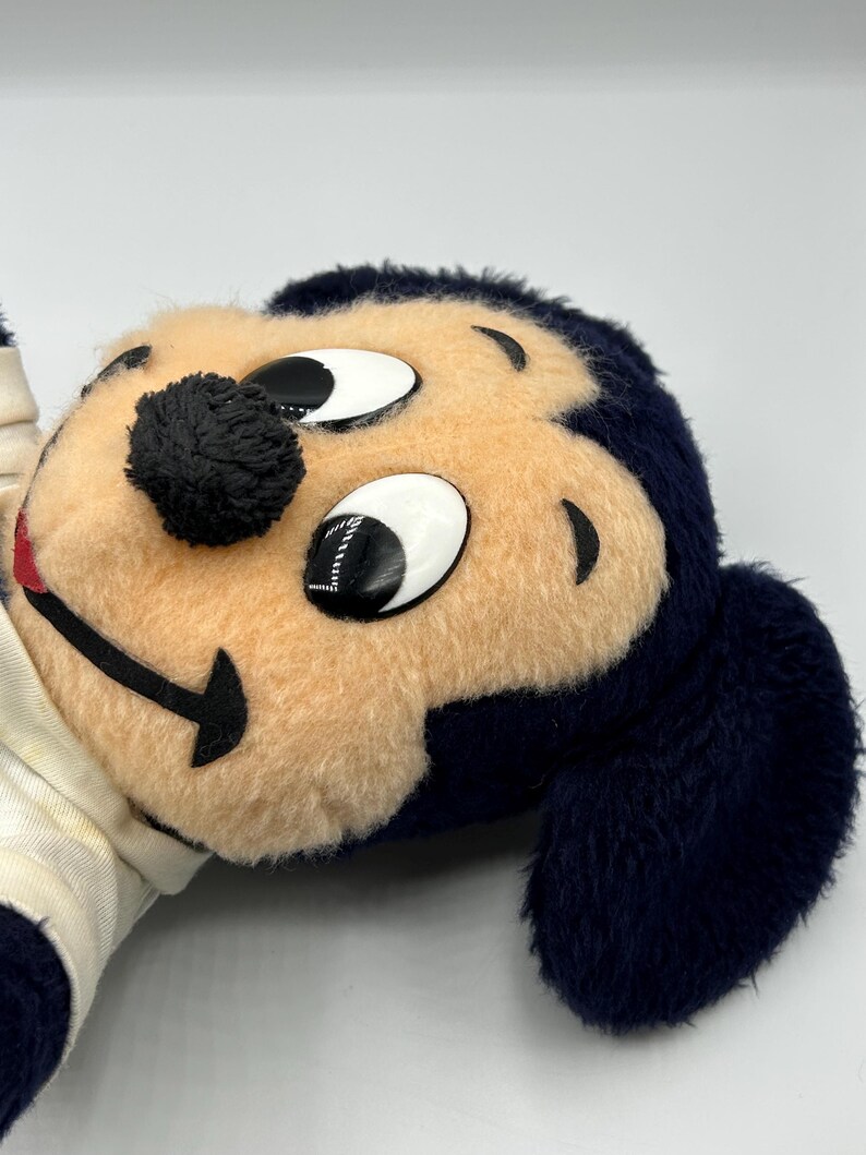 Peut inclure: Une peluche repr&eacute;sentant le visage d'un personnage de dessin anim&eacute;, avec un visage beige, des yeux noirs et un nez noir. Le personnage a des poils bleu fonc&eacute; autour du visage et des oreilles. Le jouet est partiellement visible, avec une chemise blanche et cr&egrave;me.