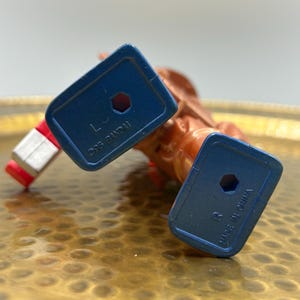 K&ouml;nnte beinhalten: Nahaufnahme einer Vintage-Actionfigur, haupts&auml;chlich orange und rot, mit blauen F&uuml;&szlig;en. Auf den F&uuml;&szlig;en steht "&copy;95 BANDAI" und "MADE IN CHINA". Die Figur liegt auf einer goldfarbenen, strukturierten Oberfl&auml;che.