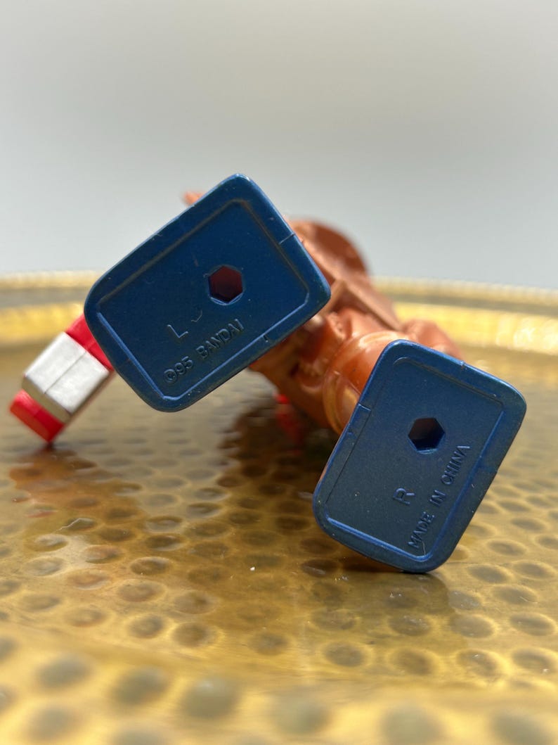 K&ouml;nnte beinhalten: Nahaufnahme einer Vintage-Actionfigur, haupts&auml;chlich orange und rot, mit blauen F&uuml;&szlig;en. Auf den F&uuml;&szlig;en steht "&copy;95 BANDAI" und "MADE IN CHINA". Die Figur liegt auf einer goldfarbenen, strukturierten Oberfl&auml;che.