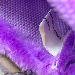 May include: Close-up of a plush, purple fabric with a textured pattern. The fabric has a soft, fuzzy texture and is trimmed with a fluffy, faux fur. A white tag with the words "POLYESTER FIBERS" is visible.