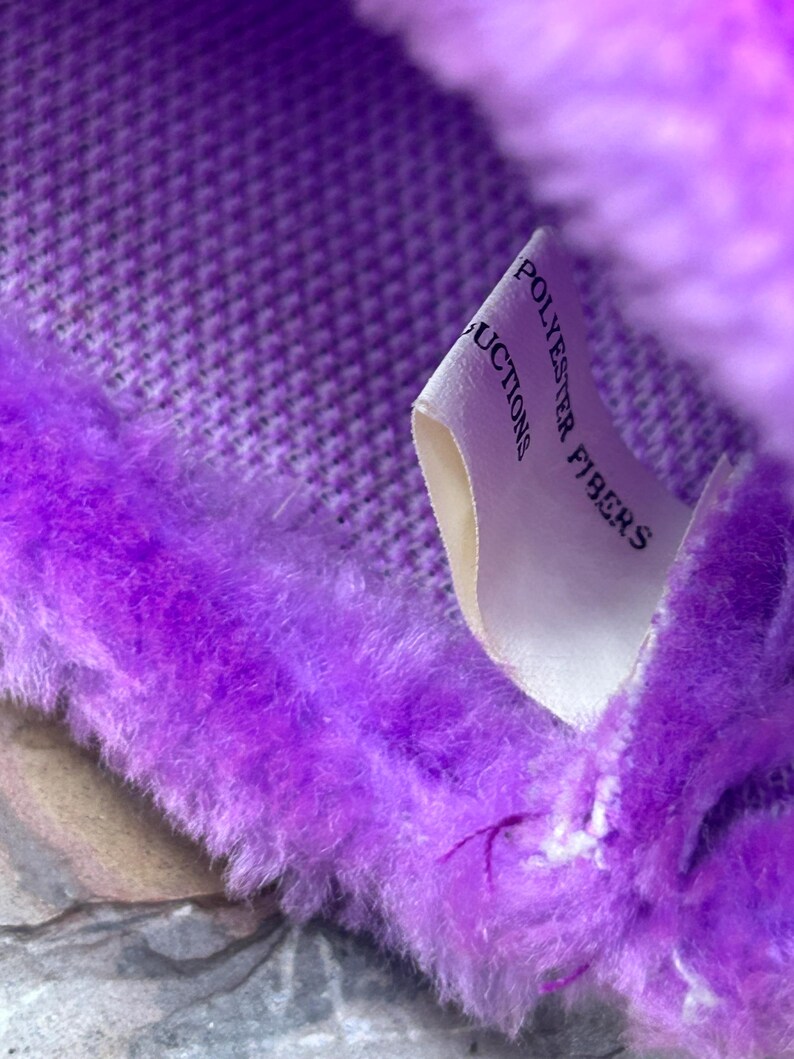 May include: Close-up of a plush, purple fabric with a textured pattern. The fabric has a soft, fuzzy texture and is trimmed with a fluffy, faux fur. A white tag with the words "POLYESTER FIBERS" is visible.