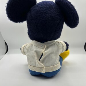 Peut inclure: Une peluche vintage de Mickey Mouse, vue de dos. La peluche pr&eacute;sente de grandes oreilles bleu fonc&eacute;, une t&ecirc;te assortie et une chemise blanche &agrave; deux boutons. Le jouet porte un pantalon bleu et des chaussures jaunes. La peluche est assise.