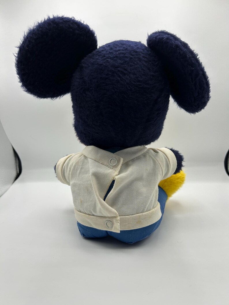 Peut inclure: Une peluche vintage de Mickey Mouse, vue de dos. La peluche pr&eacute;sente de grandes oreilles bleu fonc&eacute;, une t&ecirc;te assortie et une chemise blanche &agrave; deux boutons. Le jouet porte un pantalon bleu et des chaussures jaunes. La peluche est assise.