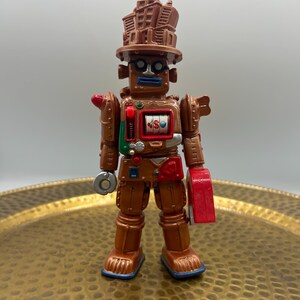K&ouml;nnte beinhalten: Ein brauner Spielzeugroboter mit einem geb&auml;udeartigen Hut, runden Gl&auml;sern und einem Spielautomaten-Design auf der Brust. Der Roboter hat eine rote Hand und einen silbernen kreisf&ouml;rmigen Gegenstand in der anderen Hand. Das Spielzeug steht auf einem goldfarbenen Tablett.