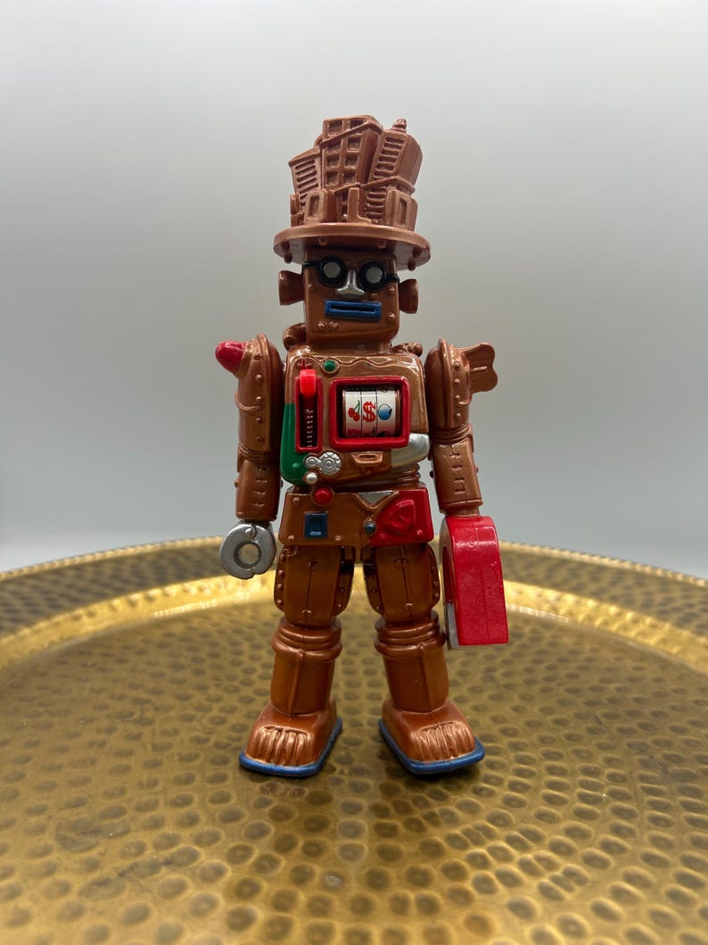 K&ouml;nnte beinhalten: Ein brauner Spielzeugroboter mit einem geb&auml;udeartigen Hut, runden Gl&auml;sern und einem Spielautomaten-Design auf der Brust. Der Roboter hat eine rote Hand und einen silbernen kreisf&ouml;rmigen Gegenstand in der anderen Hand. Das Spielzeug steht auf einem goldfarbenen Tablett.