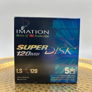 Peut inclure: Une boîte noire contenant cinq disques Imation SuperDisk 120MB LS-120. La face avant présente le logo Imation, le texte SuperDisk 120MB et un graphique d'une main tenant une baguette de chef d'orchestre. Elle inclut aussi Garantie à vie.